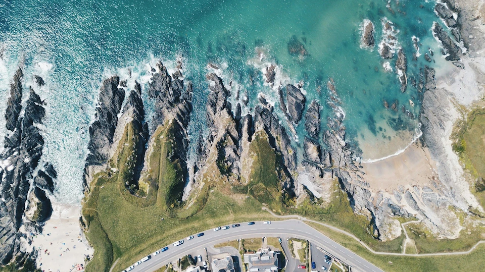 Aerial view of a rugged UK coastline with turquoise sea, jagged rocks and small sandy coves beside a clifftop road lined with houses