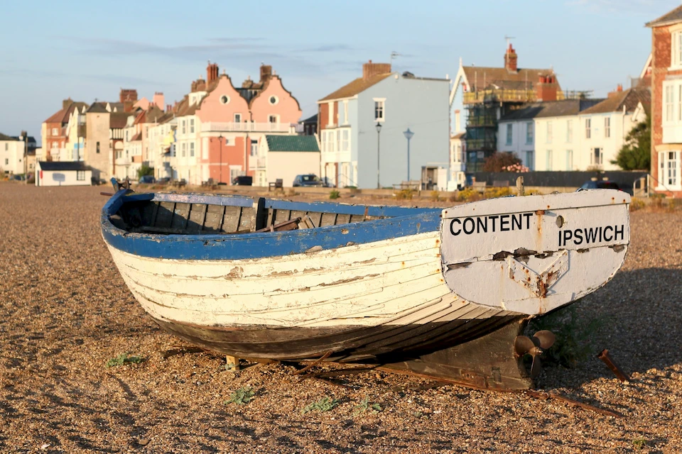 A weathered blue-and-white wooden fishing boat on the shingle beach at Aldeburgh, Suffolk, with pastel seafront houses behind