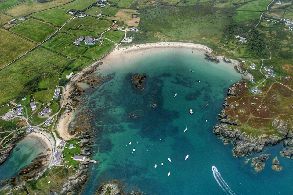 Aerial view of a sheltered sandy bay with moored boats, turquoise water and patchwork green farmland on the Welsh coast